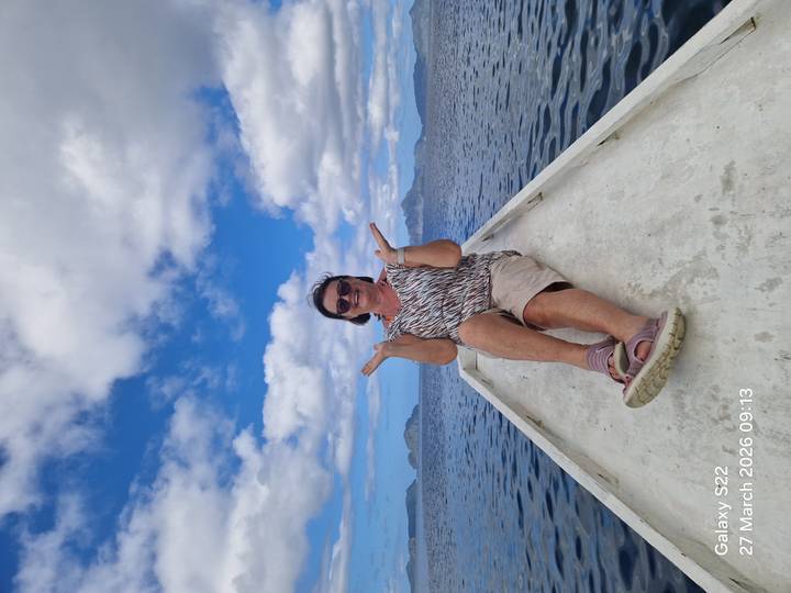 Woman posing playfully on the bow of a small boat with islands and blue sky behind; timestamp overlay on image.