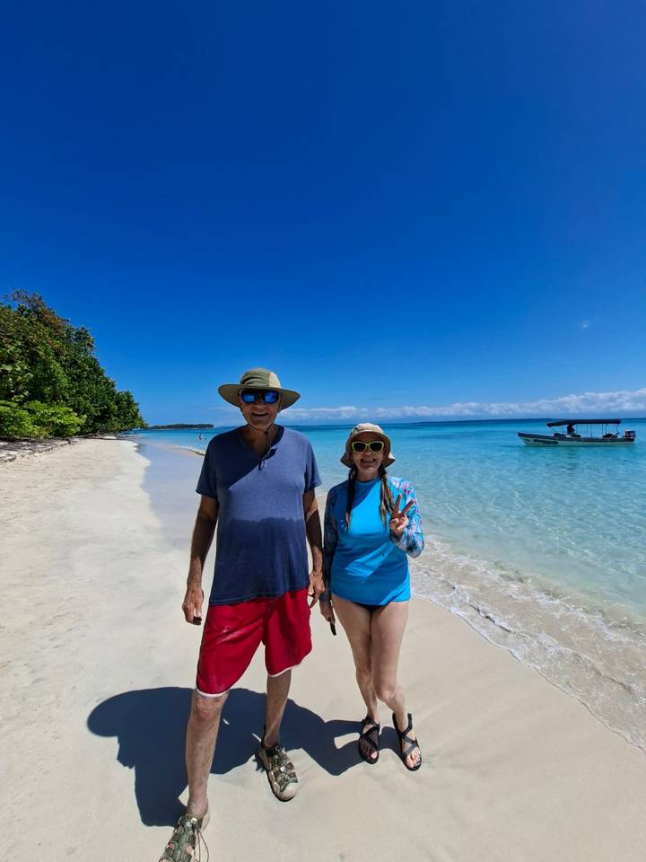 Smiling couple standing on white-sand tropical beach with clear turquoise water and small boat offshore