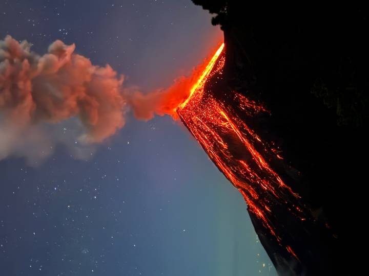 Nighttime long-exposure of an erupting volcano with glowing lava rivers and a plume of ash beneath a starry sky.
