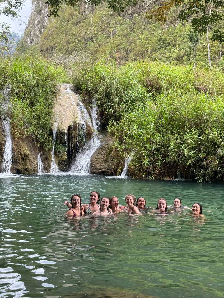 Nine travelers smile while swimming in a turquoise pool beneath a small waterfall and lush greenery.