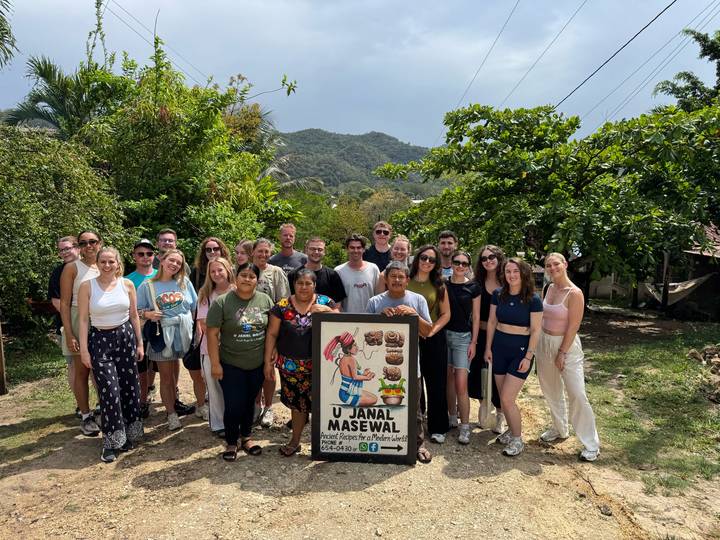 Large tour group poses with a community signboard against a backdrop of green hills and forest.