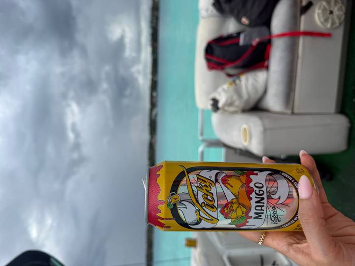 Hand holds a mango beer can on a boat deck overlooking striking turquoise lagoon waters beneath grey skies.