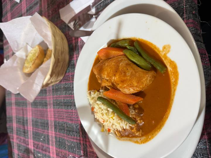 Plate of chicken in rich red sauce with rice, carrots, green beans and bread basket on a checked tablecloth.