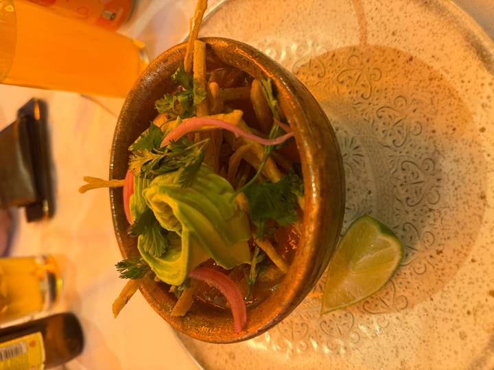 Earthen bowl of hearty soup topped with avocado slices, herbs and pickled onions under warm lighting.