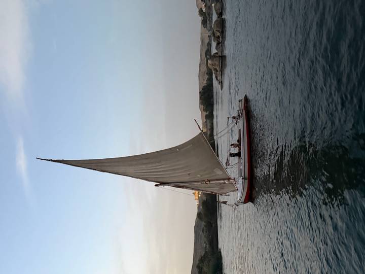 Traditional felucca sailboat glides along calm river waters at dusk under a pale sky.
