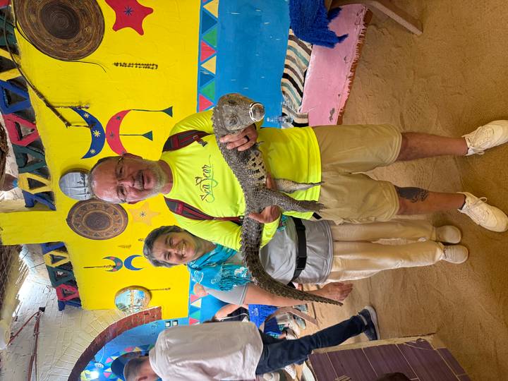 Smiling couple poses indoors holding a small crocodile in front of brightly painted Nubian artwork.