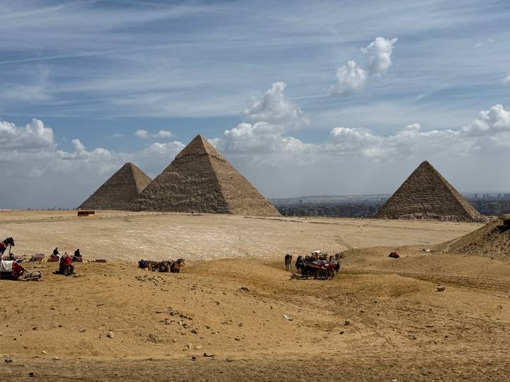 Panoramic view of the three Great Pyramids of Giza rising from the desert plateau under scattered clouds.