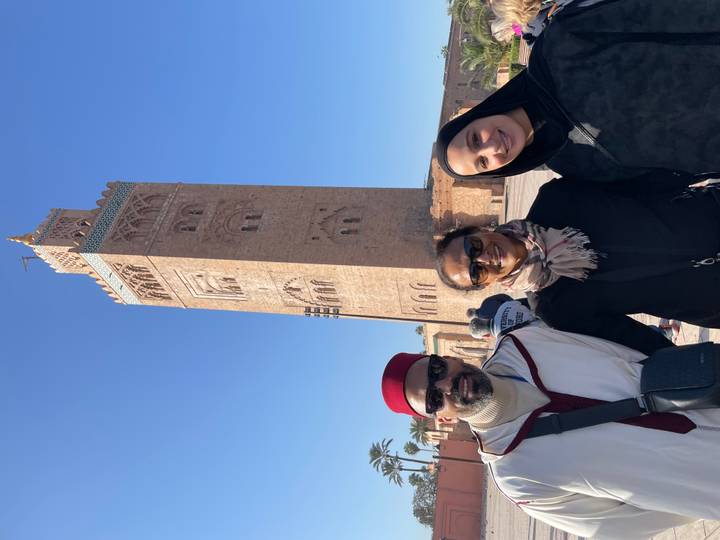 Guide and two tourists stand before the tall minaret of Koutoubia Mosque under a clear blue sky.