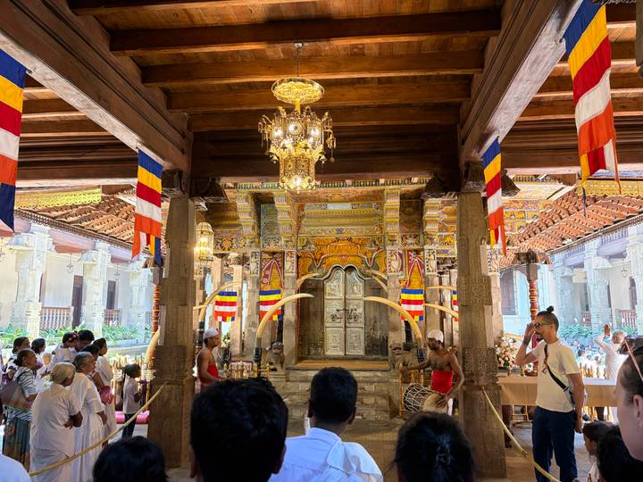 Ornate interior of a Buddhist temple with colorful flags, chandeliers, tusks and worshippers gathered for a ceremony.