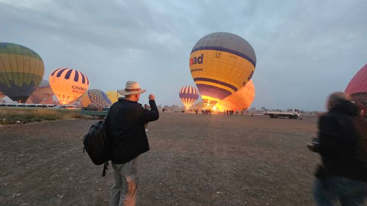 Several colorful hot-air balloons being inflated at dawn while visitors watch on the dusty launch field.