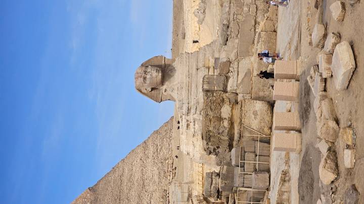 Iconic Great Sphinx with pyramid backdrop under clear blue sky, small tourists at its base.