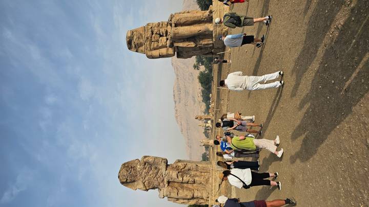 Visitors standing before the towering twin statues of the Colossi of Memnon with desert mountains behind.