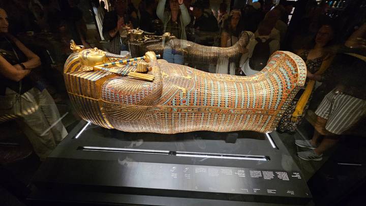 Golden, intricately decorated sarcophagus displayed in a glass case at a museum with onlookers.