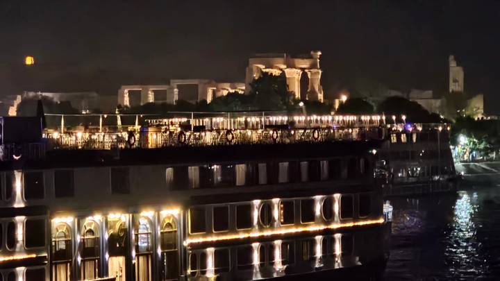 Night river scene with bright cruise ship lights and silhouette of temple ruins behind.