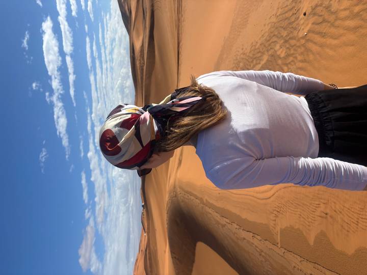 Woman with patterned headscarf stands gazing over rolling orange sand dunes under blue sky.