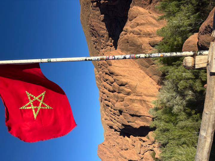 Moroccan flag waving on a pole against dramatic reddish rock formations and blue sky.