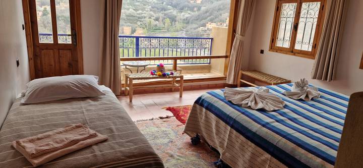 Cozy guesthouse room with twin beds, striped blankets and balcony overlooking green mountain valley.