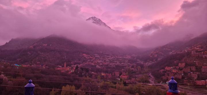 Pink sunset light bathes a mist-filled Atlas mountain peak and the small village nestled in the green valley below.