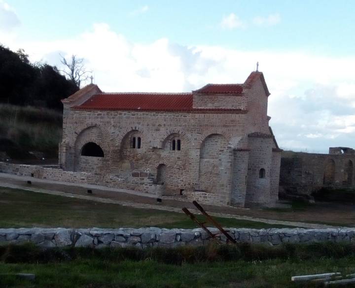 Stone church ruins with red-tiled roof sit on a grassy site under a cloudy sky.