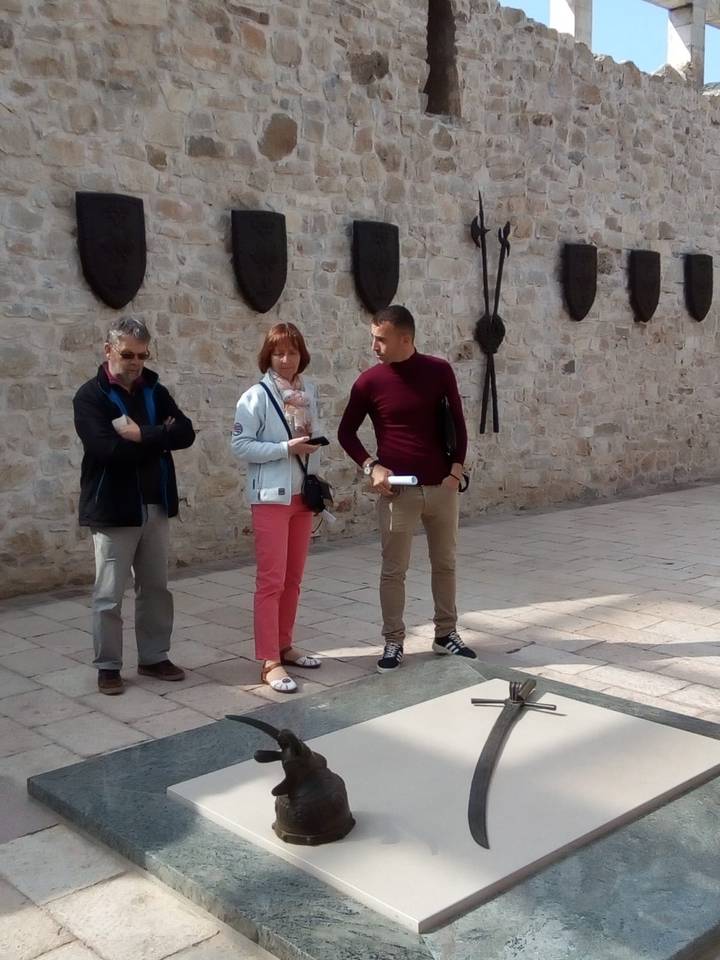 Three visitors listen to a guide beside a stone wall adorned with medieval weapons.