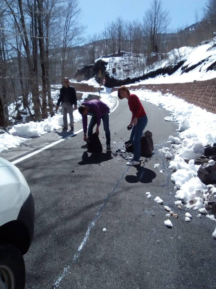 Travellers clear fallen rocks from a snow-lined mountain road on a sunny winter day.