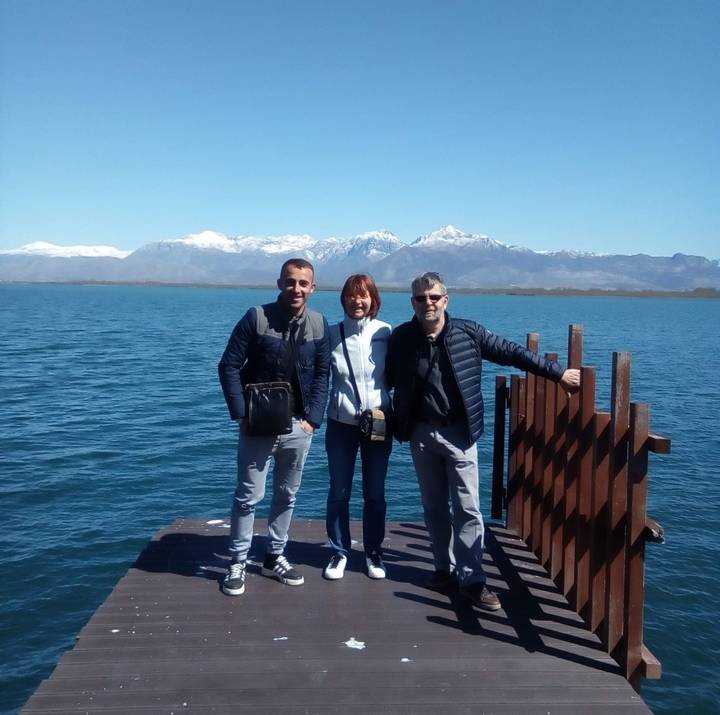 Three friends pose on a wooden pier with a blue lake and snow-capped mountains behind them.