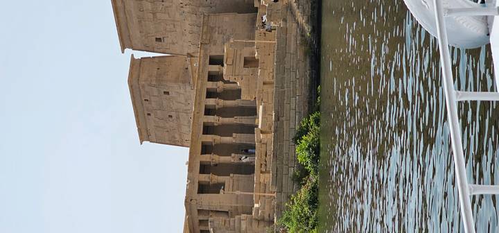 Ancient riverside temple complex viewed from a boat on calm water under clear sky