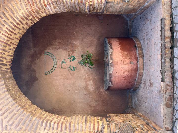 Small plant growing from round copper well beneath faded mural inside arched niche