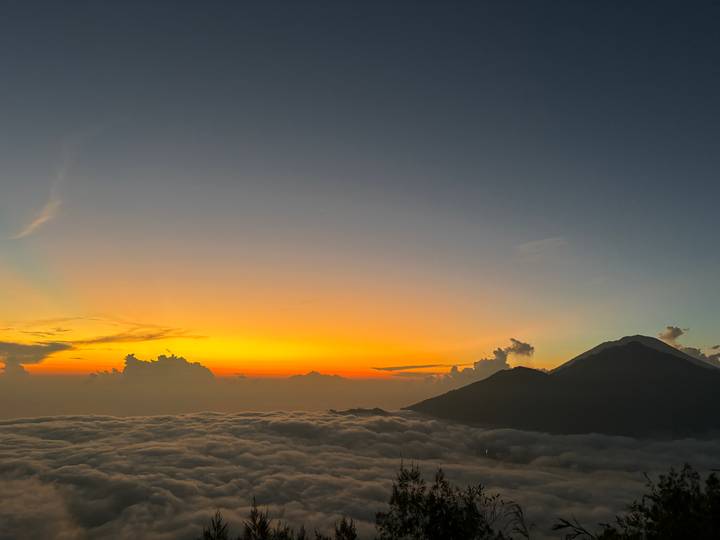 Golden sunrise spreads above clouds with a silhouetted volcano peak.