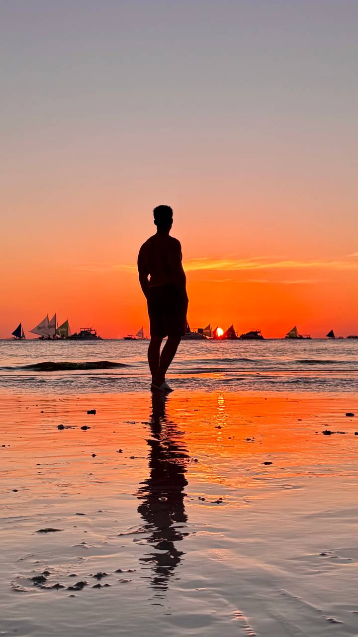 Silhouette of a man on the beach watching a vivid orange Boracay sunset with sailboats.