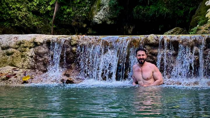 Man relaxes waist-deep in a natural pool beneath a small tropical waterfall surrounded by lush greenery.