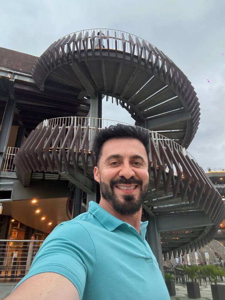 Selfie of a smiling man standing in front of a modern spiral staircase structure on a cloudy day.