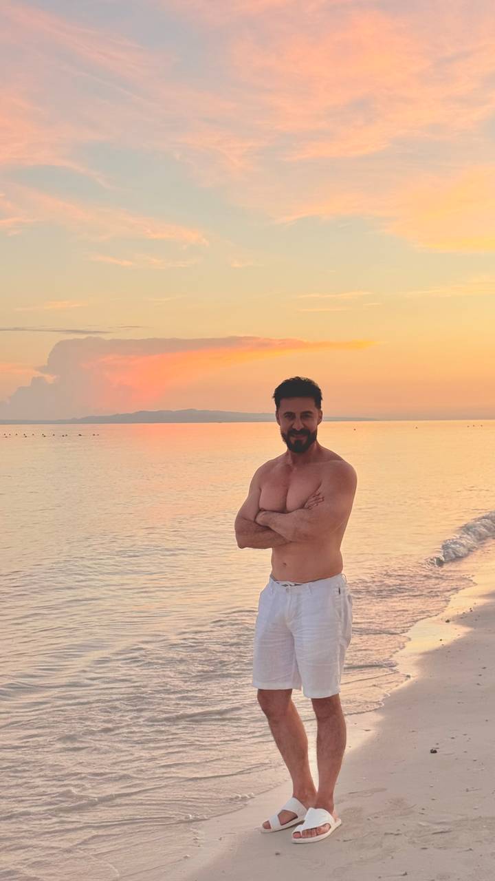 Shirtless man stands on a calm beach at colorful sunset with gentle waves and distant island silhouette.