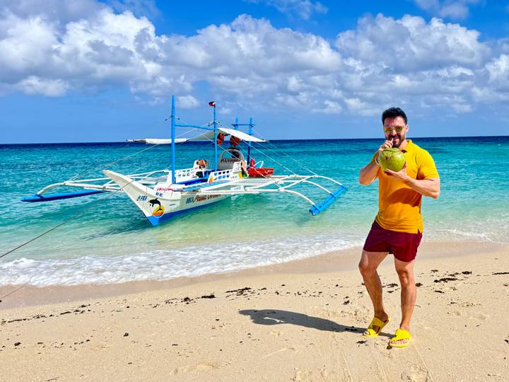 Man sips fresh coconut on white-sand beach beside turquoise sea and traditional outrigger boat under bright sun.