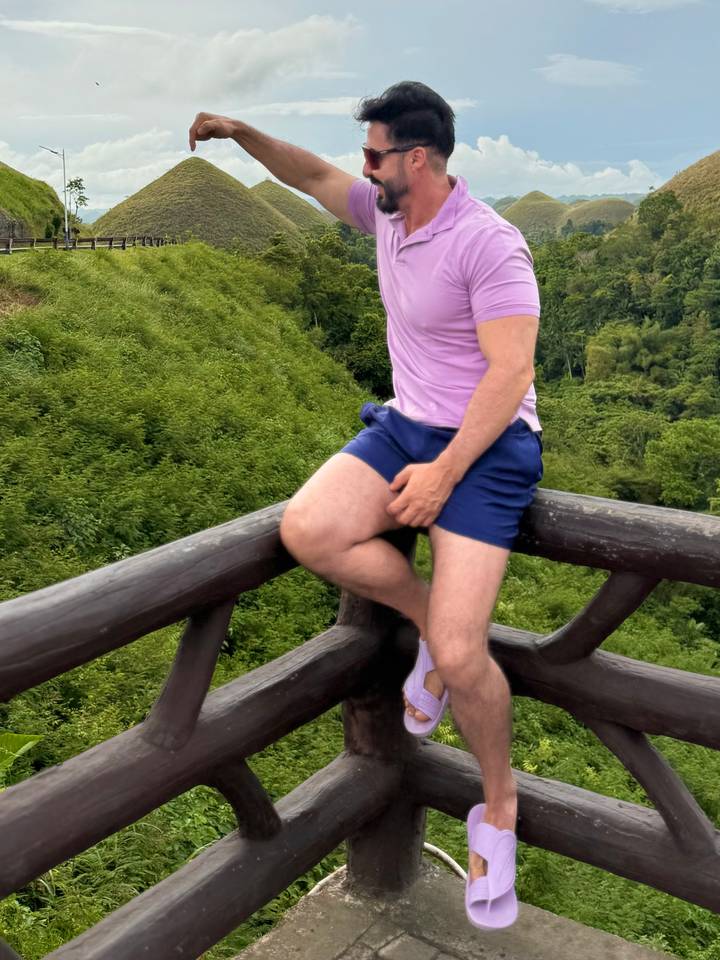 Traveler poses seated on a wooden rail overlooking lush green hills.