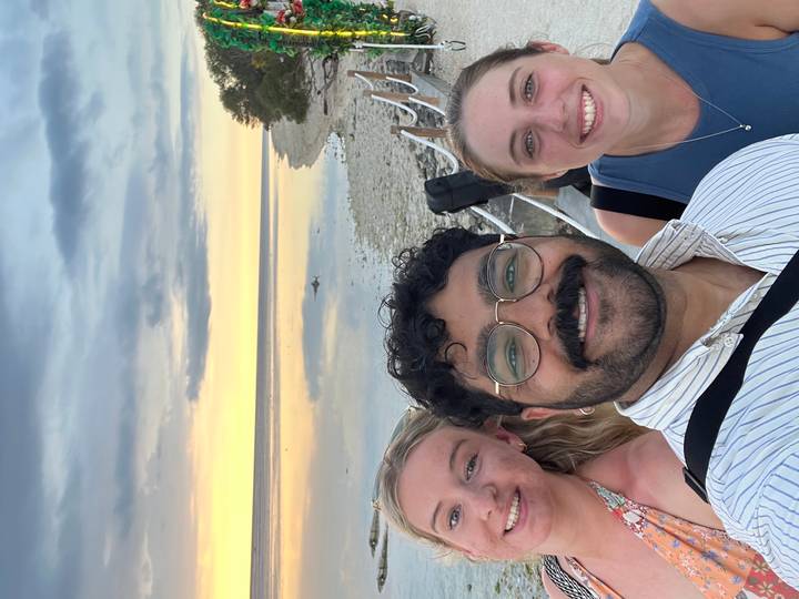 Vertical selfie of three friends smiling at serene coastal sunset with tidal flats.