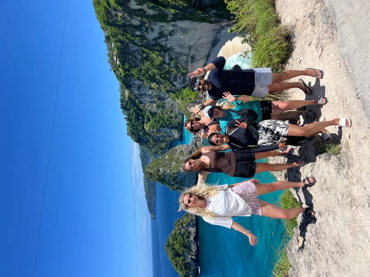 Group of hikers pose at dramatic cliffside viewpoint above turquoise bay on Nusa Penida.