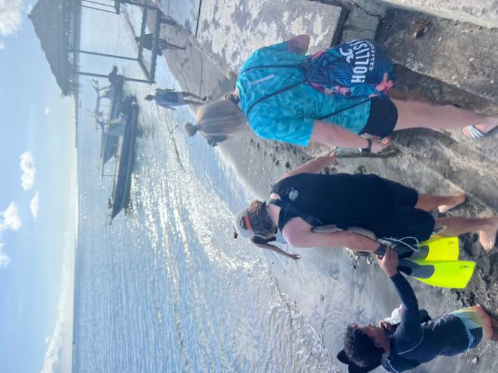 Travelers walk down sandy steps toward a small boat at the shoreline in bright light.