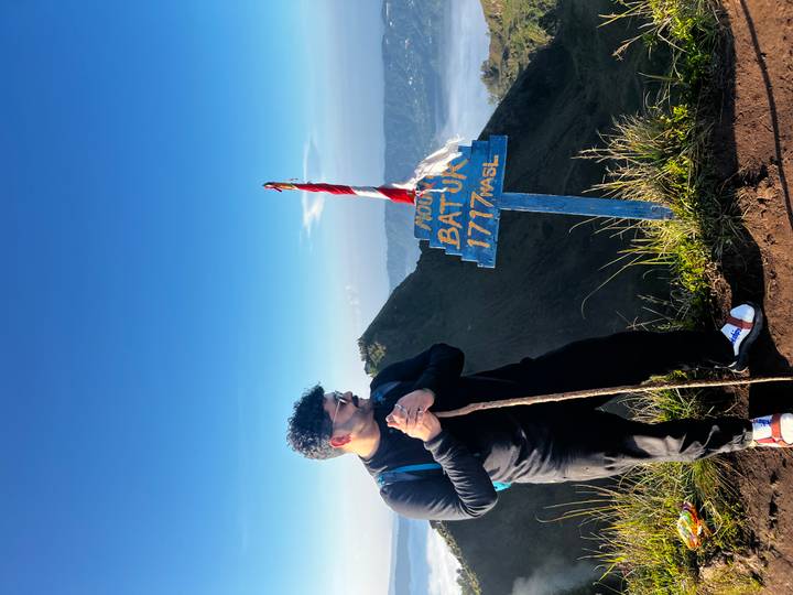 Hiker holding walking stick stands beside blue sign marking Mount Batur summit elevation.