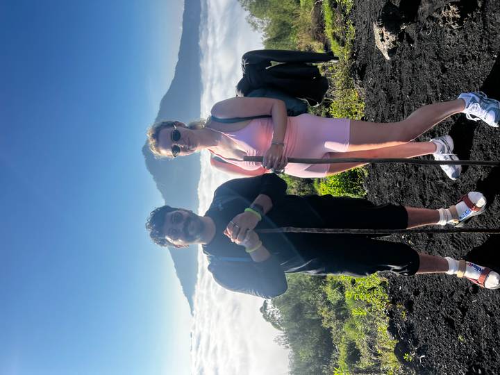 Two hikers pose on black volcanic soil with cloud-filled valley and Mount Agung behind them.