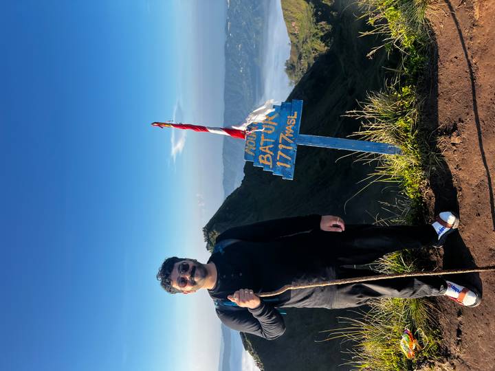 Solo hiker with staff stands proudly next to Mount Batur summit sign under clear blue sky.
