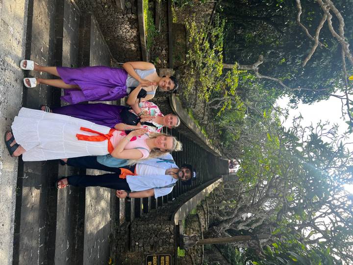 Travelers dressed in sarongs pose on stone steps leading to a Balinese temple shaded by frangipani trees.
