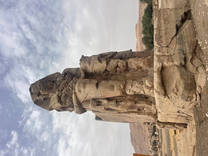 Massive seated Colossus statue carved from sandstone stands under partly cloudy skies in Luxor.