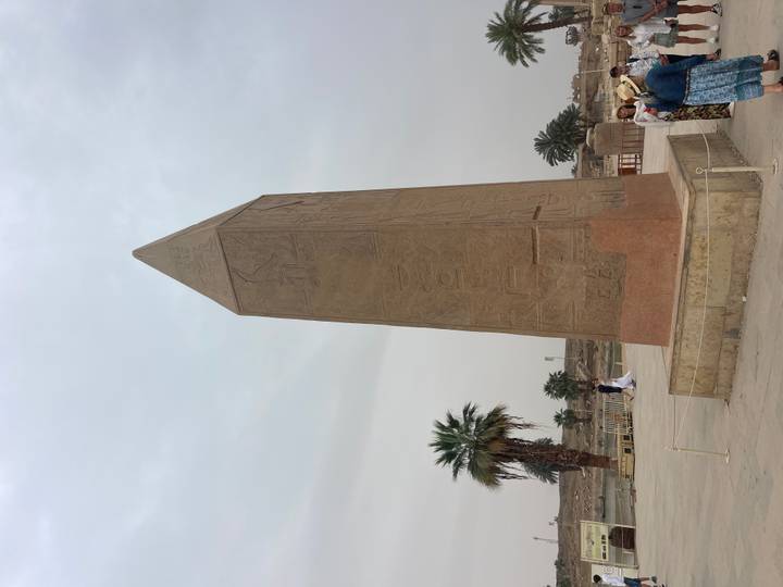 Tall ancient obelisk rises against a hazy sky with a lone palm tree nearby.