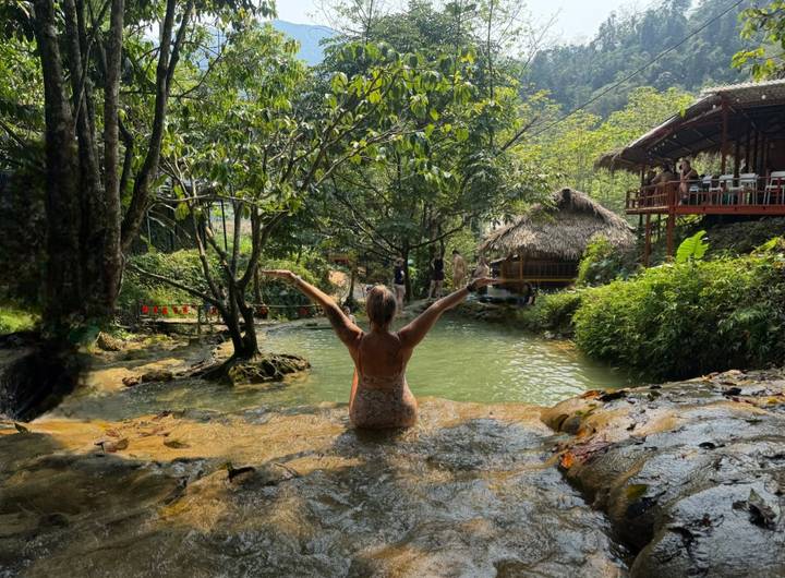 Traveler sits waist-deep in a natural spring pool surrounded by lush vegetation, arms outstretched in joy.