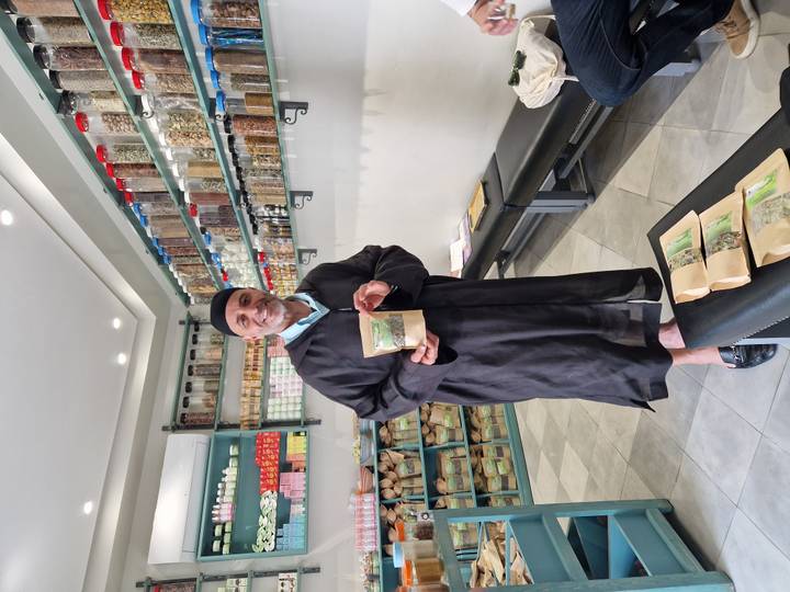 Shopkeeper in traditional robe presenting packaged spices inside an herbal store