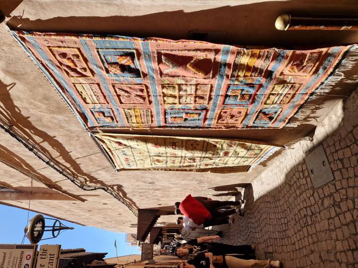 Sunlit alley with vibrant woven carpets hanging on adobe wall and pedestrians passing by