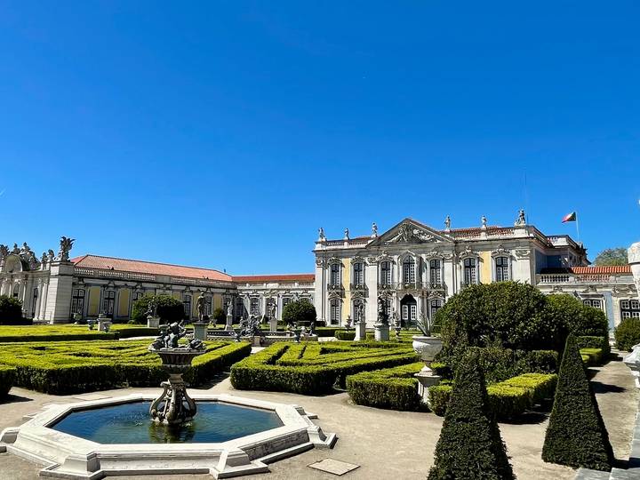 Elegant Baroque palace with manicured hedged gardens and fountains under a deep blue sky.