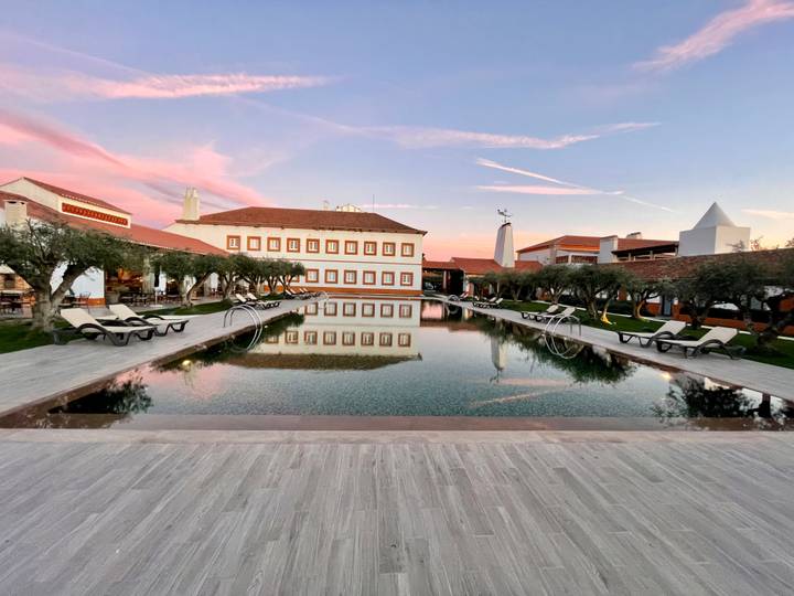 Luxurious countryside resort at sunset with symmetrical infinity pool, loungers and olive trees reflecting pastel skies.