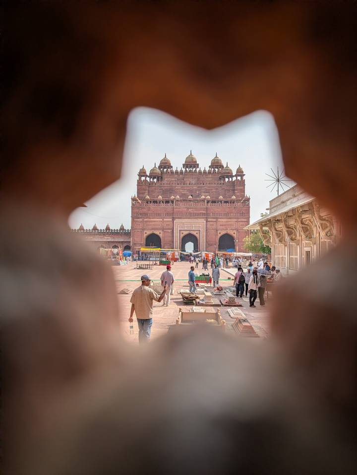 Red-sandstone Mughal gateway seen through a carved opening with market vendors and visitors in the courtyard.